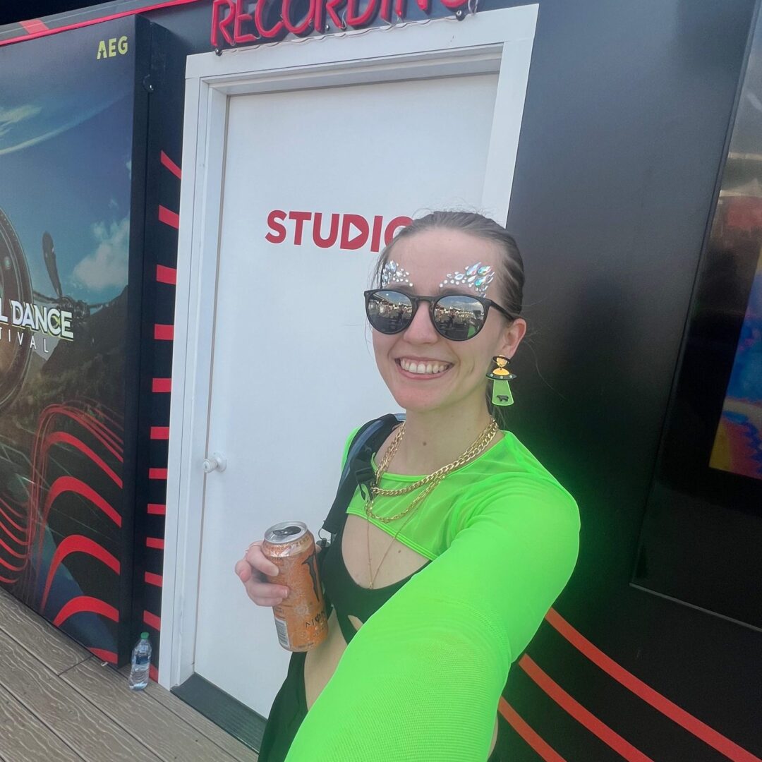 Dominique taking a selfie in front of a white door that says "Studio" with a neon sign that says "Recording" above it. She is wearing face jewels, sunglasses, and a green top. 