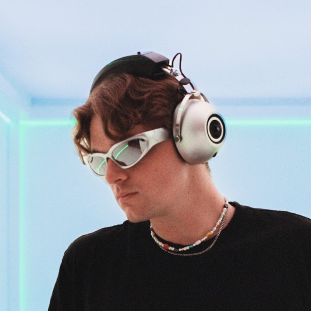 pictured in color with a light blue and retro background is the artist, veggi, who is wearing sports shades and headphones replicating a 90s techno vibe in modern day. 
